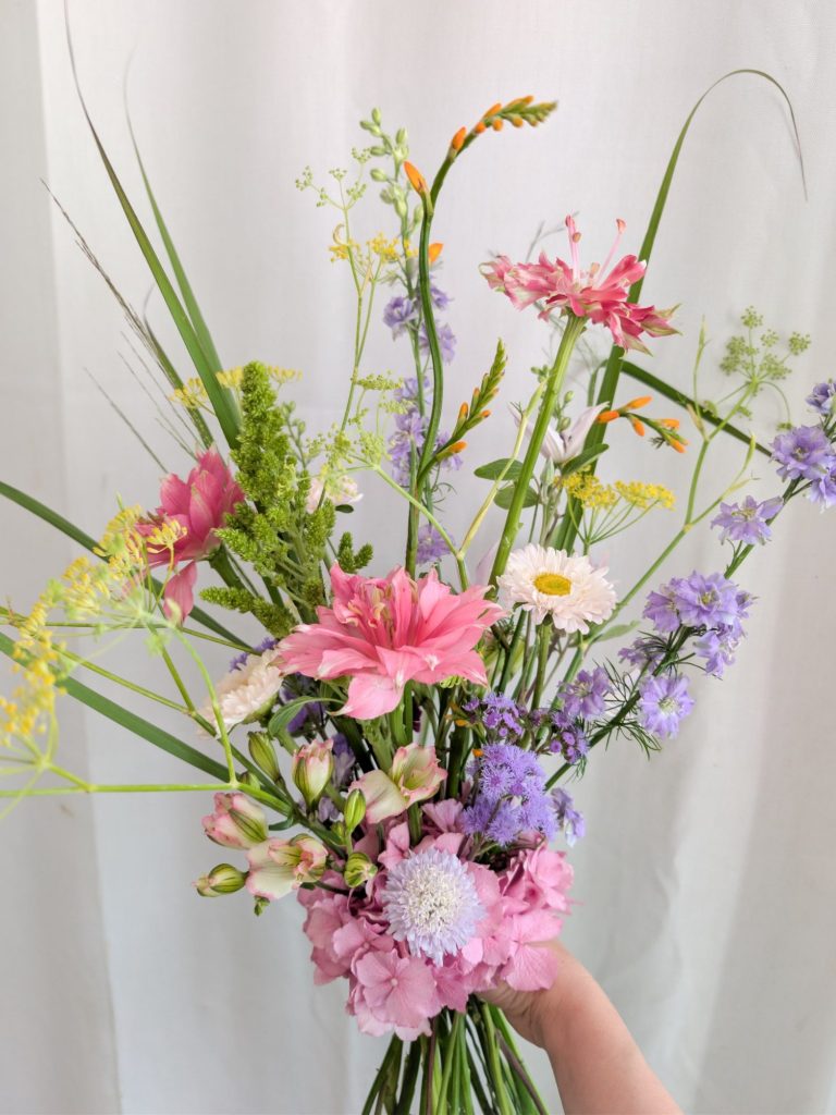 Braut hält einen bunten Blumenstrauß mit blauem, rosa und weißem Blumenschmuck.