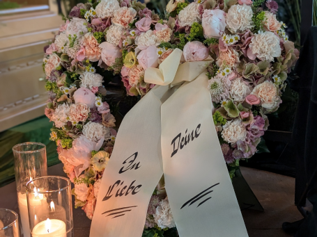 Trauerherz Ein Blütenkranz in Herzform mit einer Trauerschleife und Kerzen im Hintergrund.